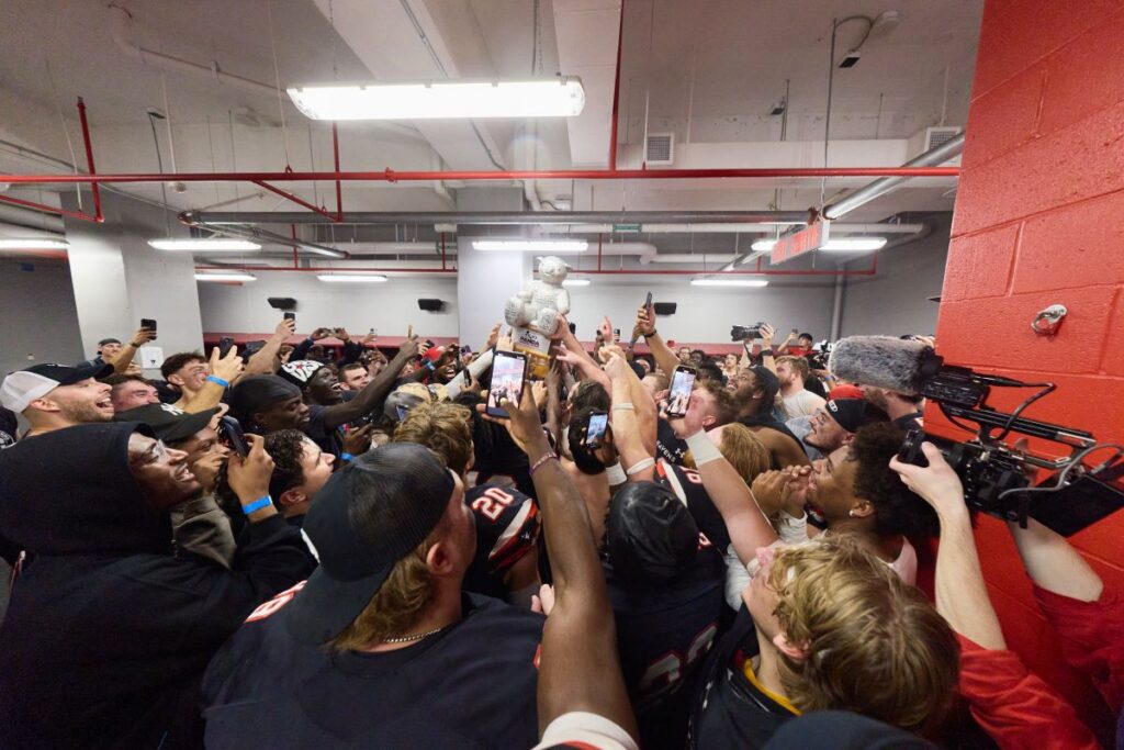 Carleton Ravens players raise the Panda Game trophy high in the locker room surrounded by cheering teammates.
