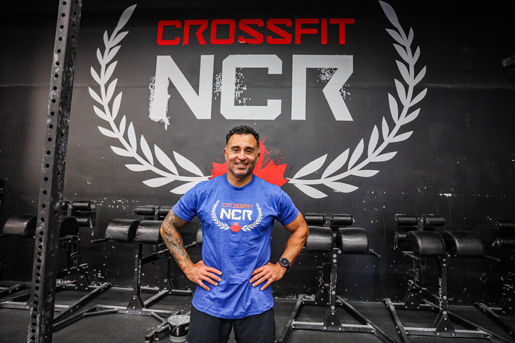 A man smiles for the camera with his hands on his hips, while standing inside a CrossFit gym.
