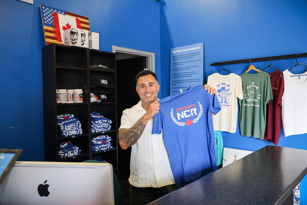 Reza Mashkoori holds up a blue CrossFit NCR T-shirt inside the gym’s merchandise area.