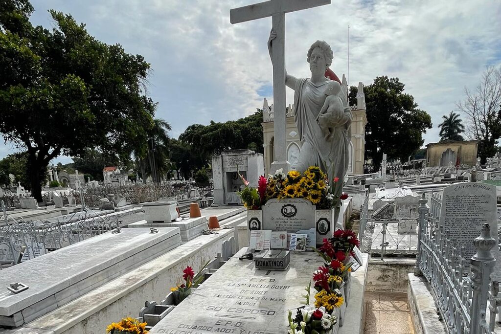 An angel holds a cross over a tomb covered in flowers, inside a large cemetery.