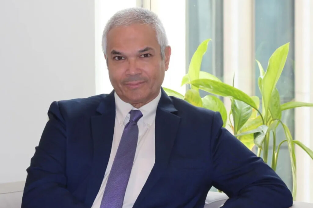 A man in a suit smiles for the camera while seated at a desk.