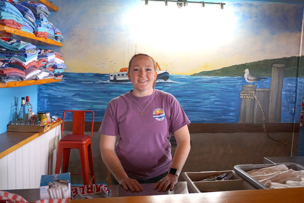 A woman with a purple shirt poses for a photo behind the counter of a restaurant.
