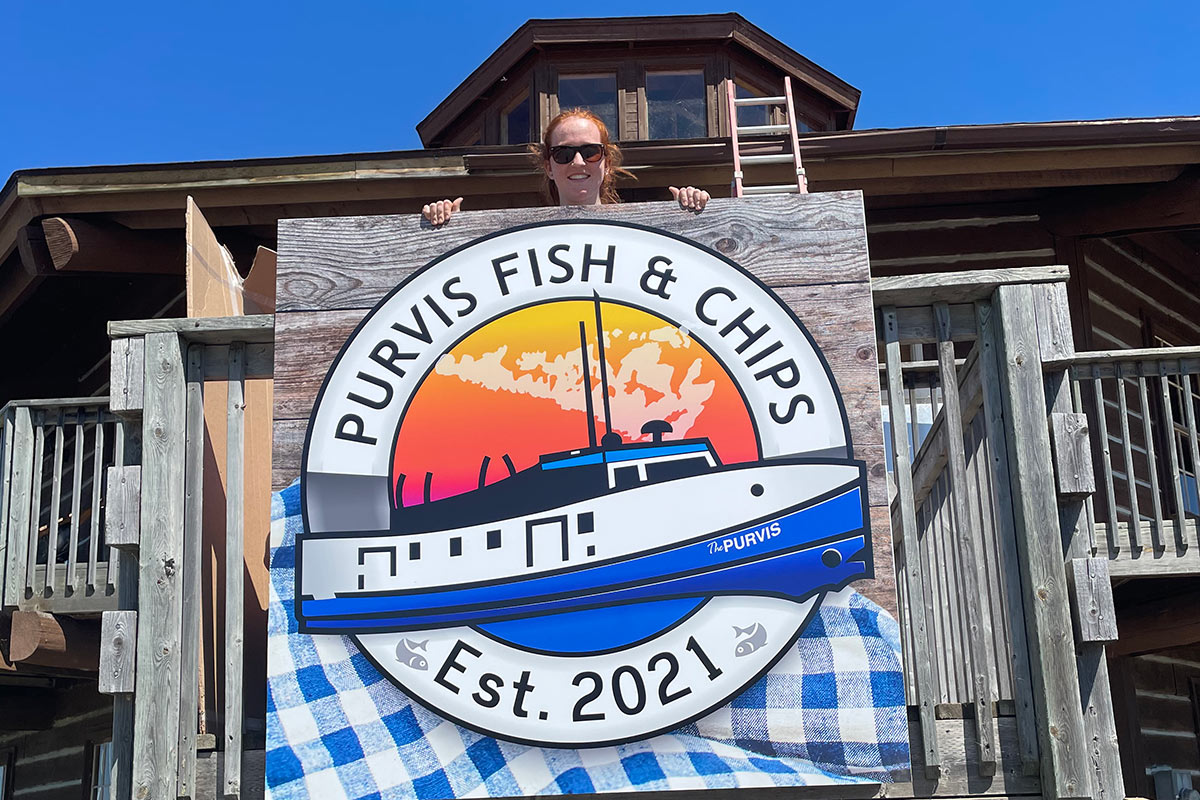 A woman wearing sunglasses stands behind a wooden sign for the restaurant Purvis Fish and Chips.