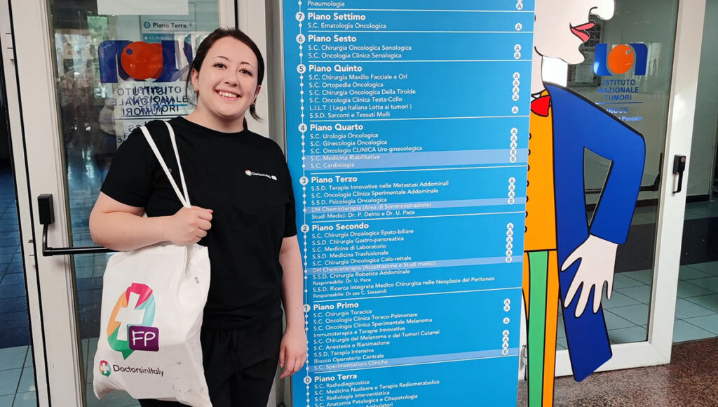 A woman wearing hospital scrubs poses for a photo next to a sign in front of an Italian hospital.