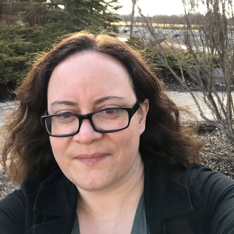 A woman with long hair and glasses takes a selfie with a river behind her.