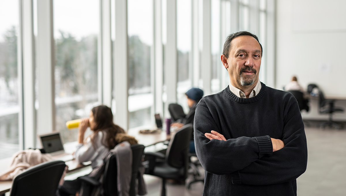 A man wearing a dark sweater over a light dress shirt poses for a photo inside a classroom, while folding his arms.