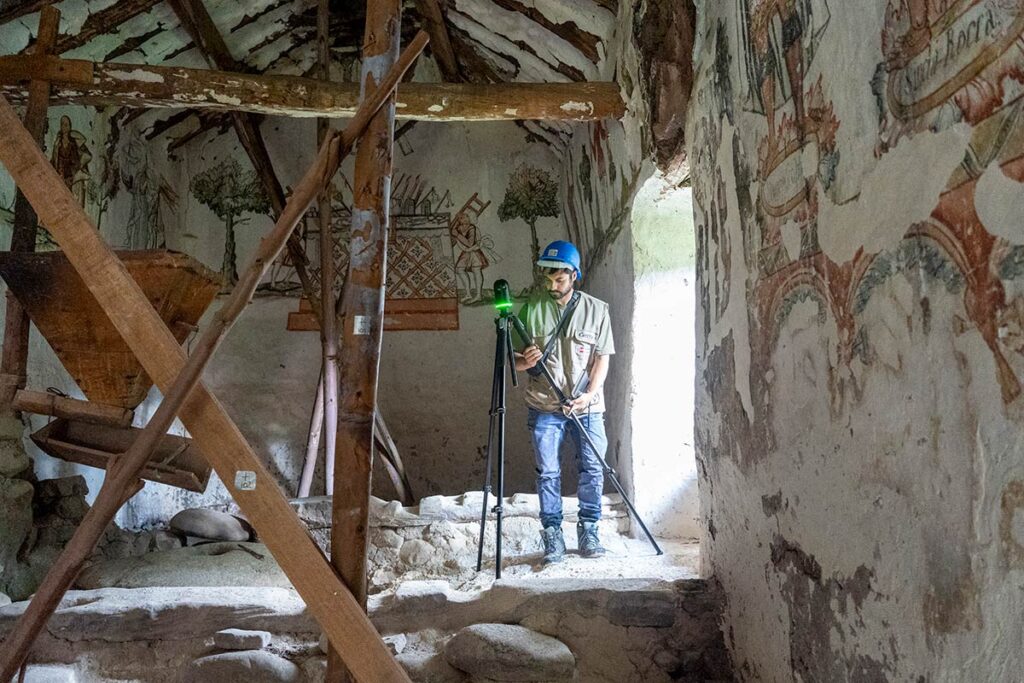 A conservation worker wearing a blue hard hat uses a laser scanner to document the interior of an old mural-covered building.