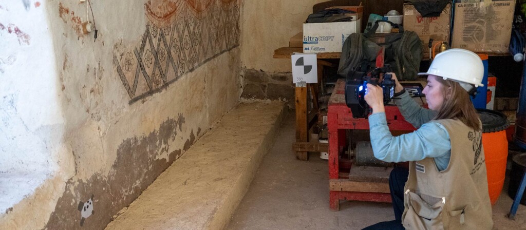 A conservation specialist wearing a hard hat photographs ancient wall paintings inside a historic building to document restoration details.