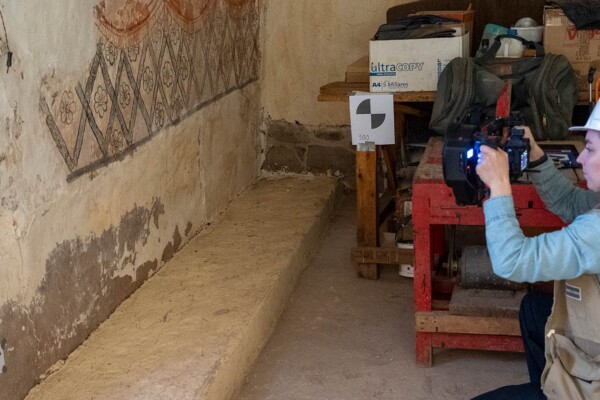 A conservation specialist wearing a hard hat photographs ancient wall paintings inside a historic building to document restoration details.