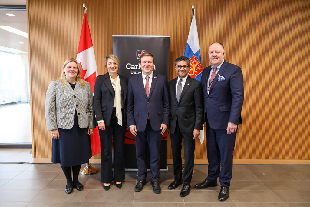 Five professionally dressed people posing for a group photo in front of a banner for Carleton University.