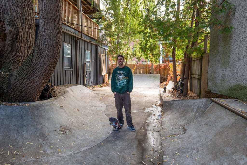 A man stands with his skateboard in a small backyard skate bowl surrounded by trees and wooden fences.