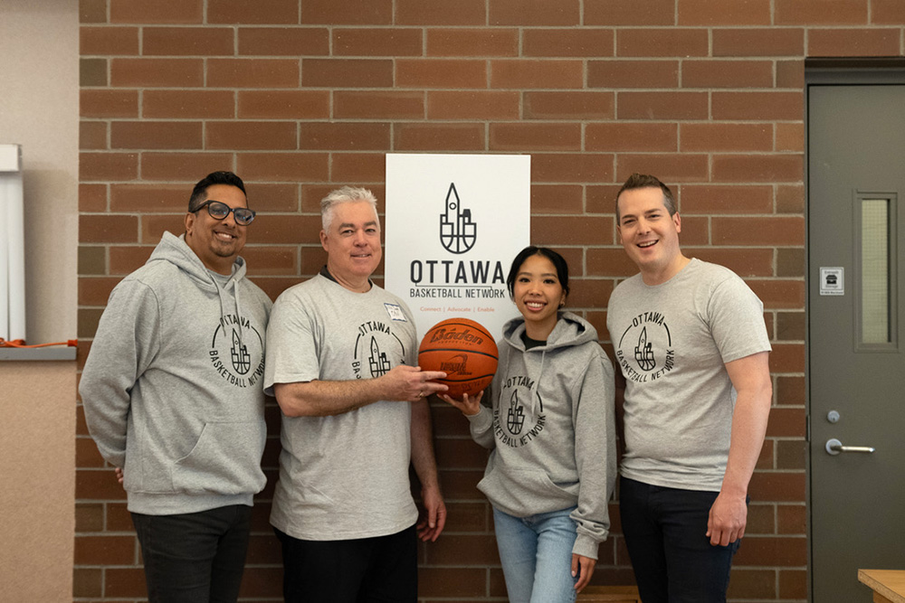 Four people wearing matching shirts pose for a photo in front of a brick wall, while one holds a basketball.