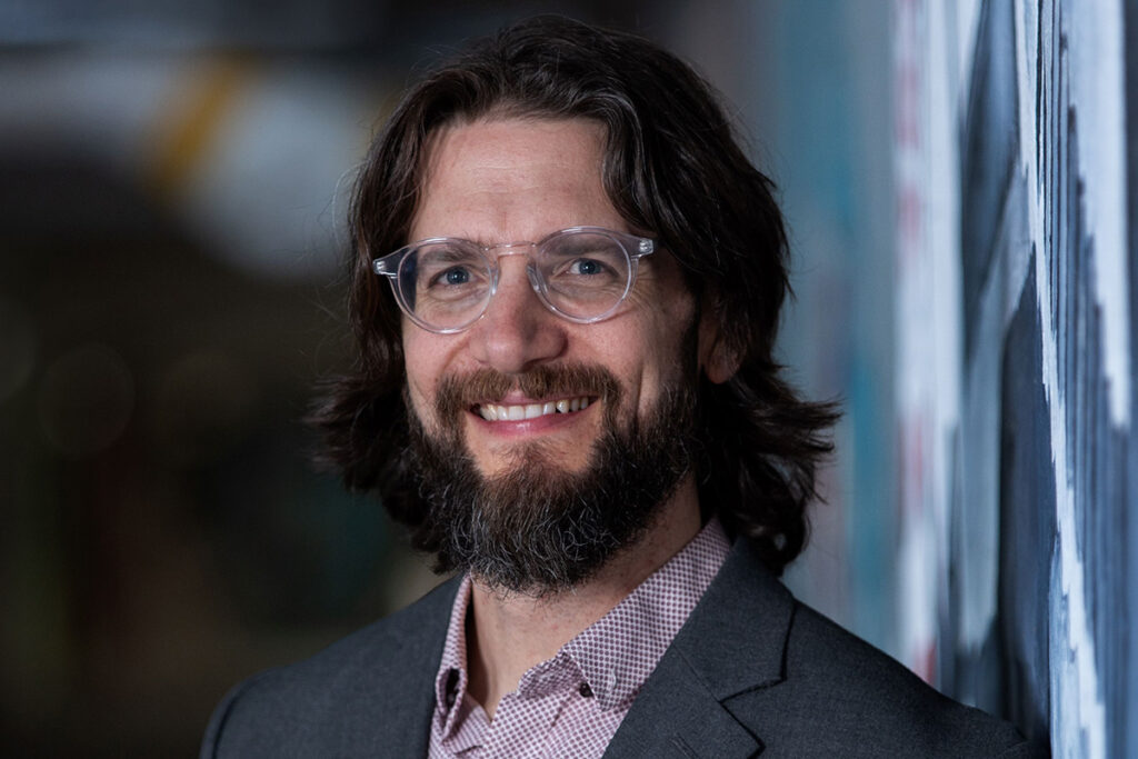 A man with a beard and glasses poses for a photo will wearing a suit.