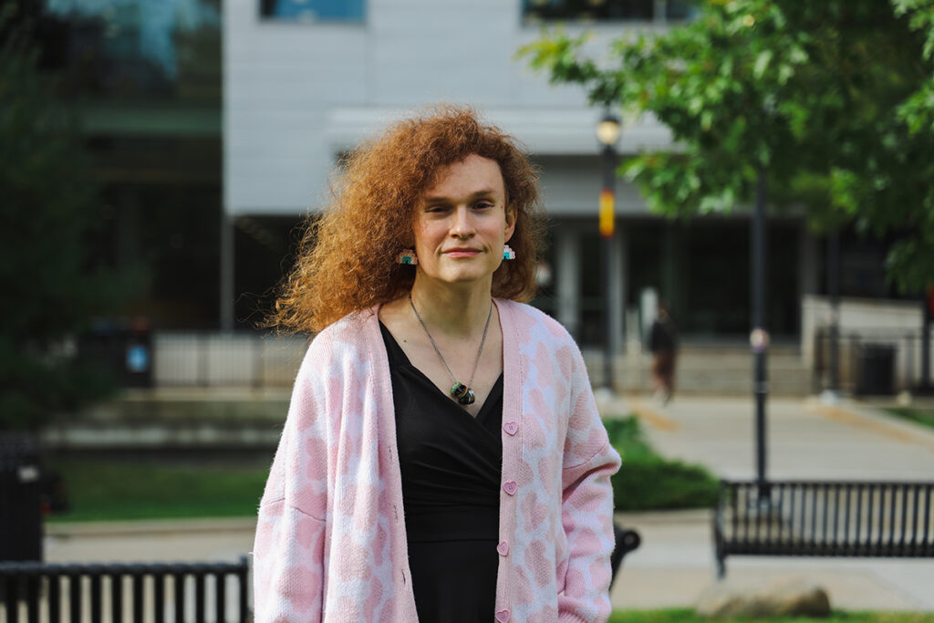 A woman with a pink cardigan and black shirt poses for a professional photo in front of a building.