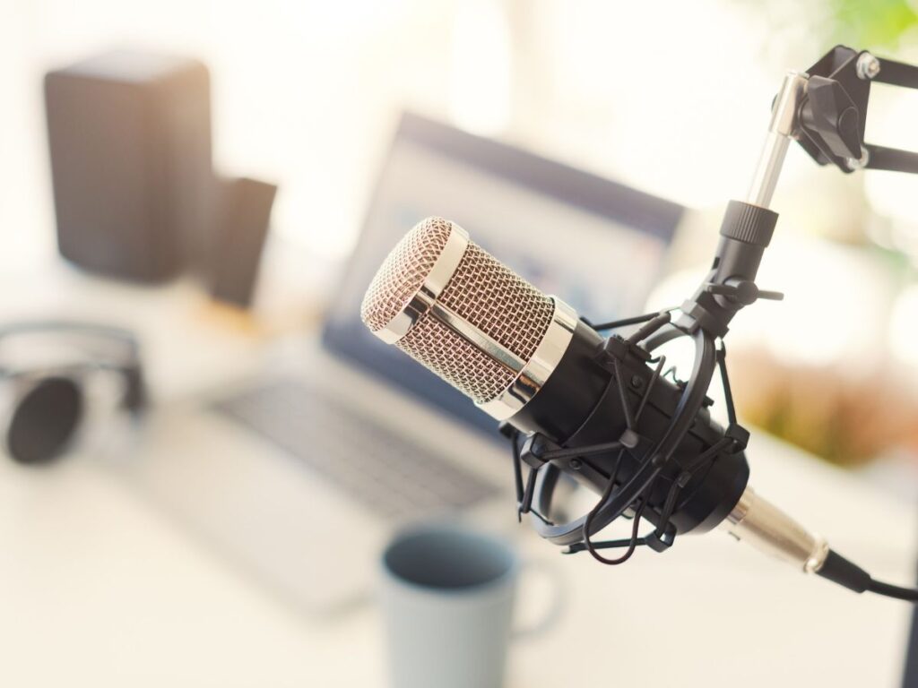 A podcast microphone is seen in front of a desk. A mug, laptop and sunglasses can be seen in the background.