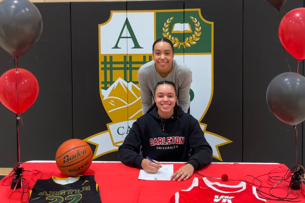Two people posing for a photo at a table surrounded by red and black balloons; one appears to be signing a document.