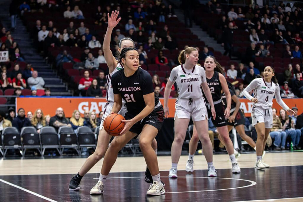 A basketball player guards the ball while a defender attempts to block her.