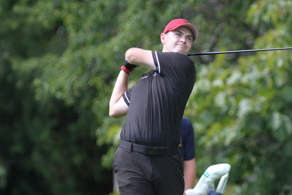 A young golfer wearing a red hat and a black shirt is captured mid-swing.