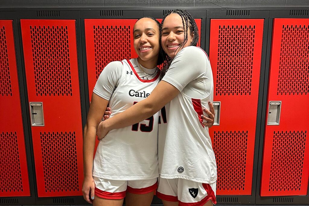 Two basketball players embracing while posing for the camera, inside a locker room.
