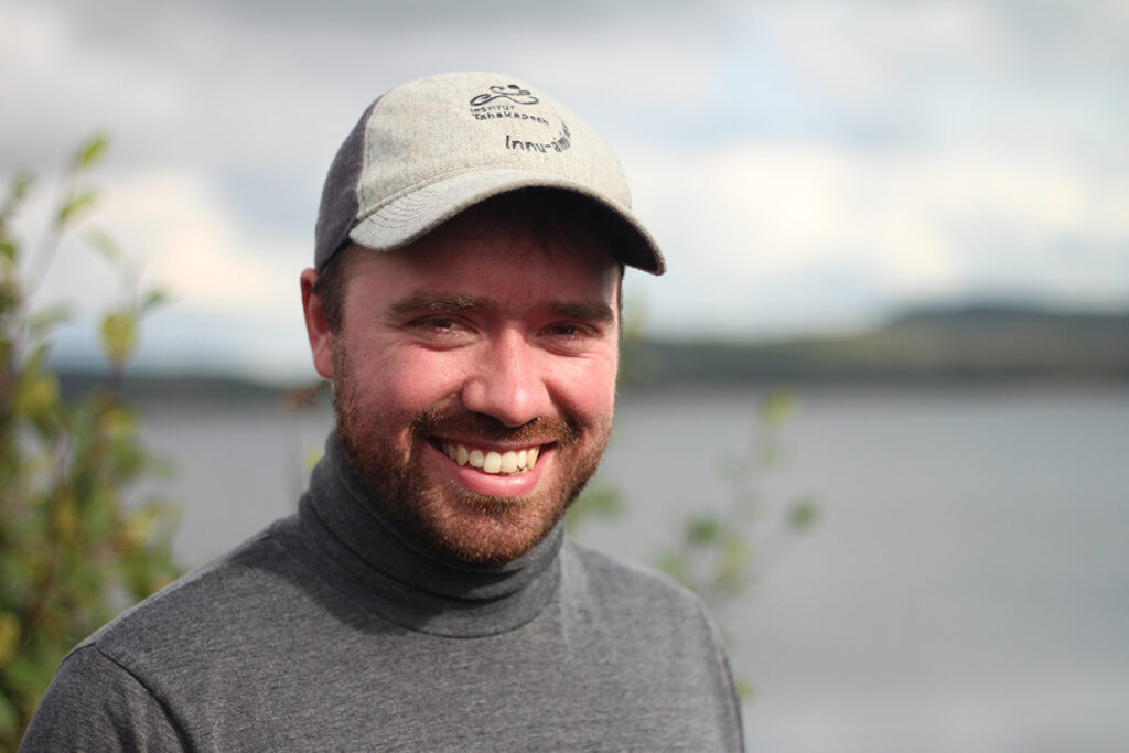 A man wearing a hat and a grey turtleneck sweater poses for a photo near a body of water.