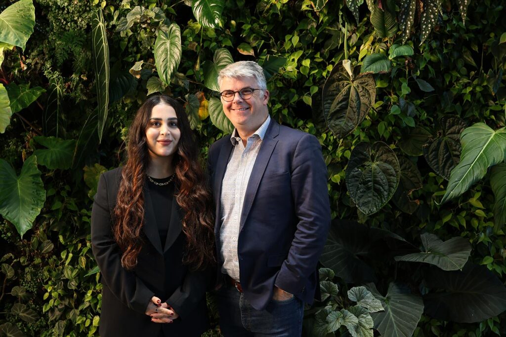 A man and a woman posing for a photo in front of a wall covered in plants.