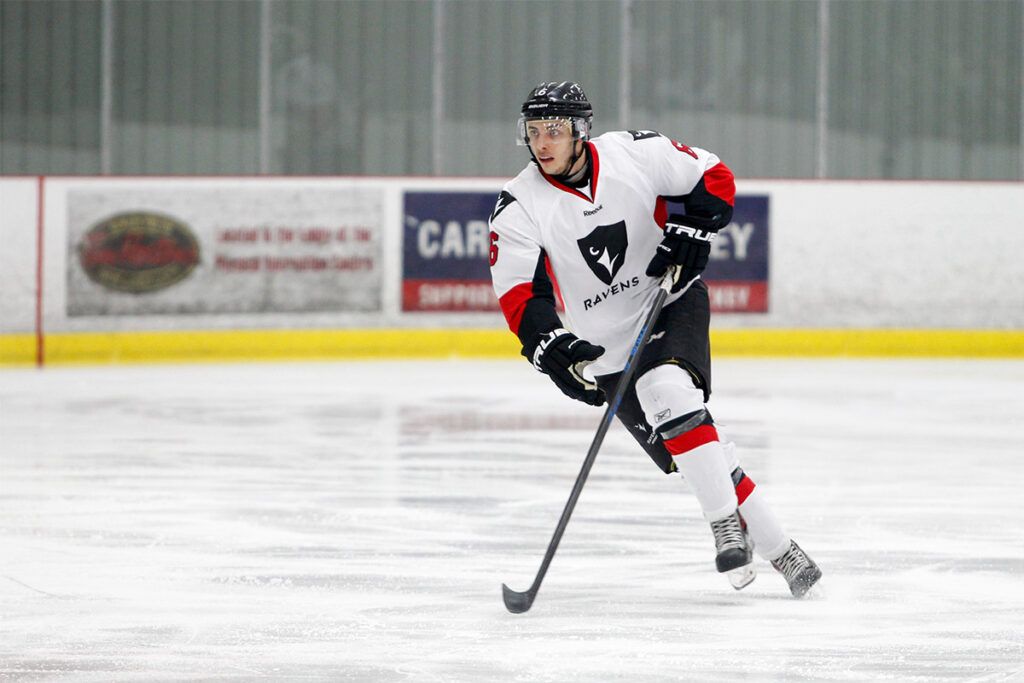 A hockey player wearing a Carleton University Ravens jersey controls the puck while skating up the ice.