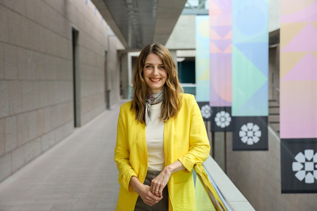 A woman wearing a yellow dress coat poses for a photo while leaning against a railing.