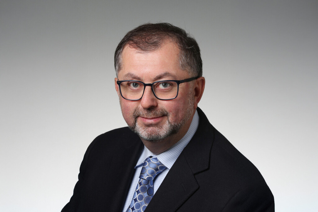 A professional headshot of a man in a dark suit, wearing glasses.