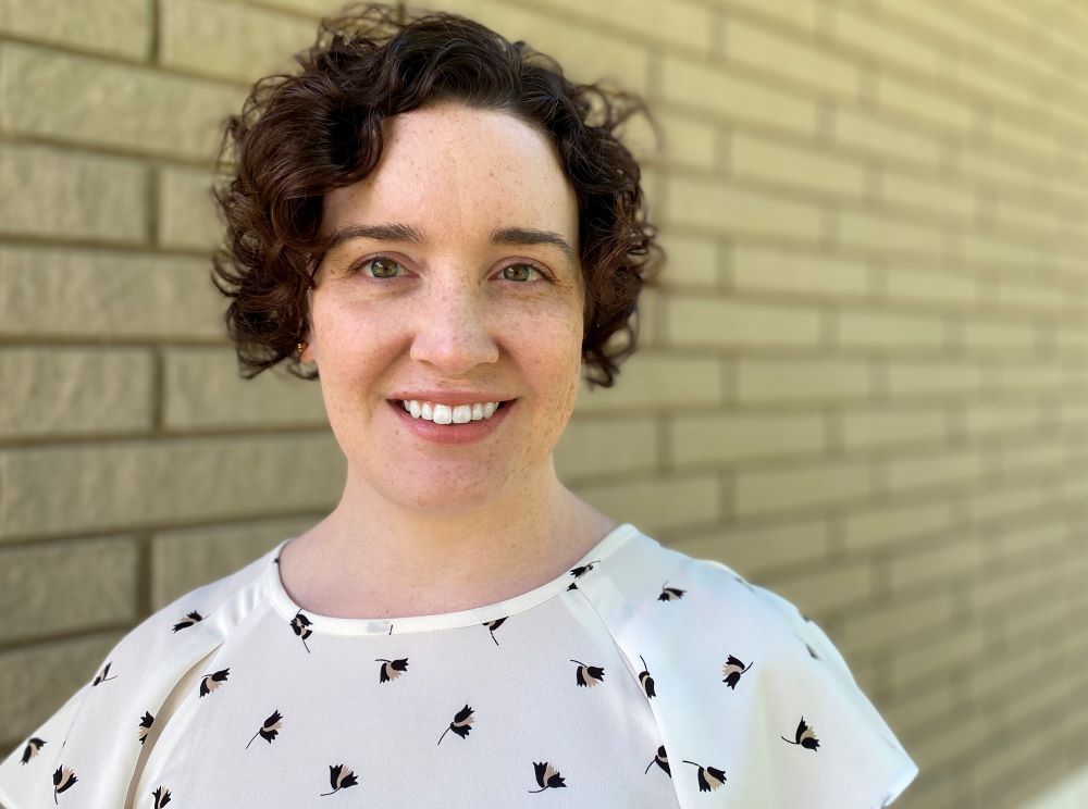 A woman wearing a white shirt smiles for the camera in front of a brick wall.