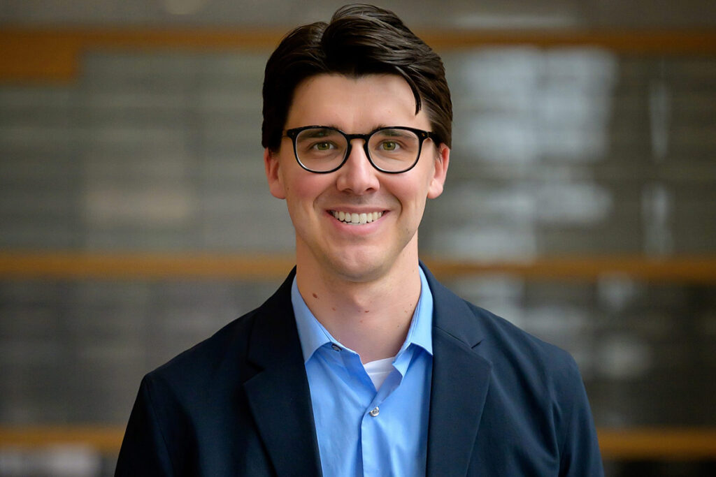 A man with short dark hair and glasses, wearing a navy blue blazer, poses for a a photo.