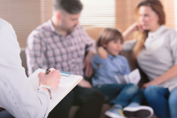 Hand of a professional family psychotherapist writing notes in front of a couple with a child in a blurred background.
