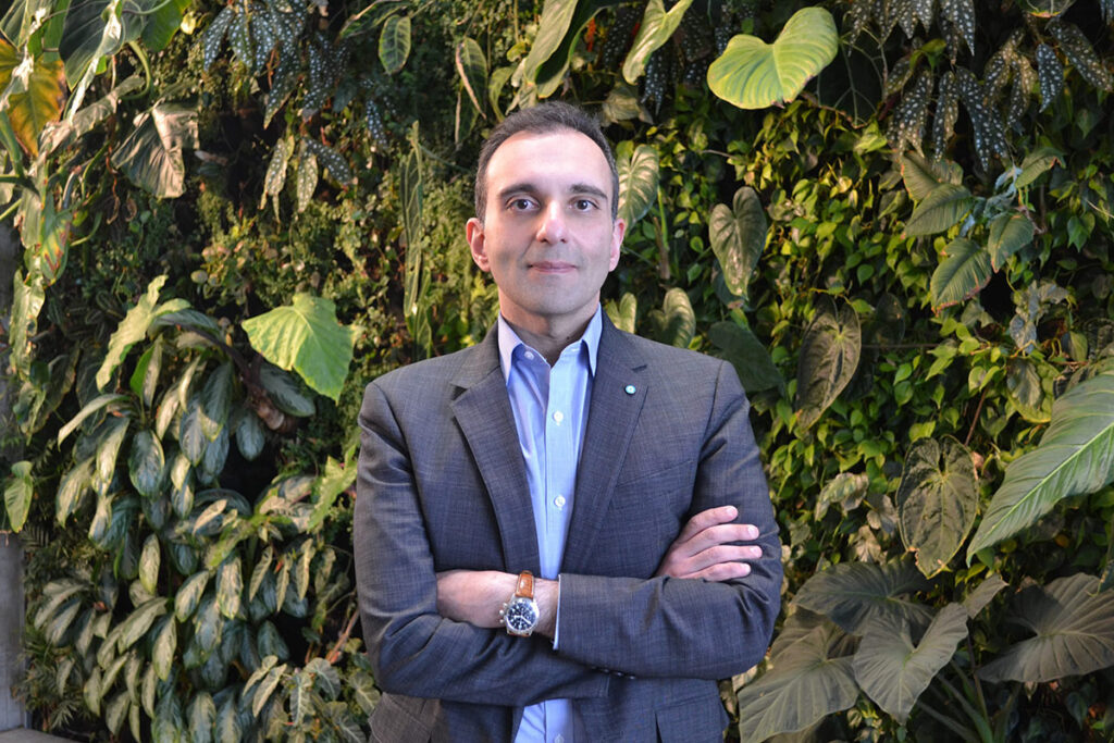 A man wearing a blue suit, with his arms crossed, poses for the camera in front of a wall covered in plants.