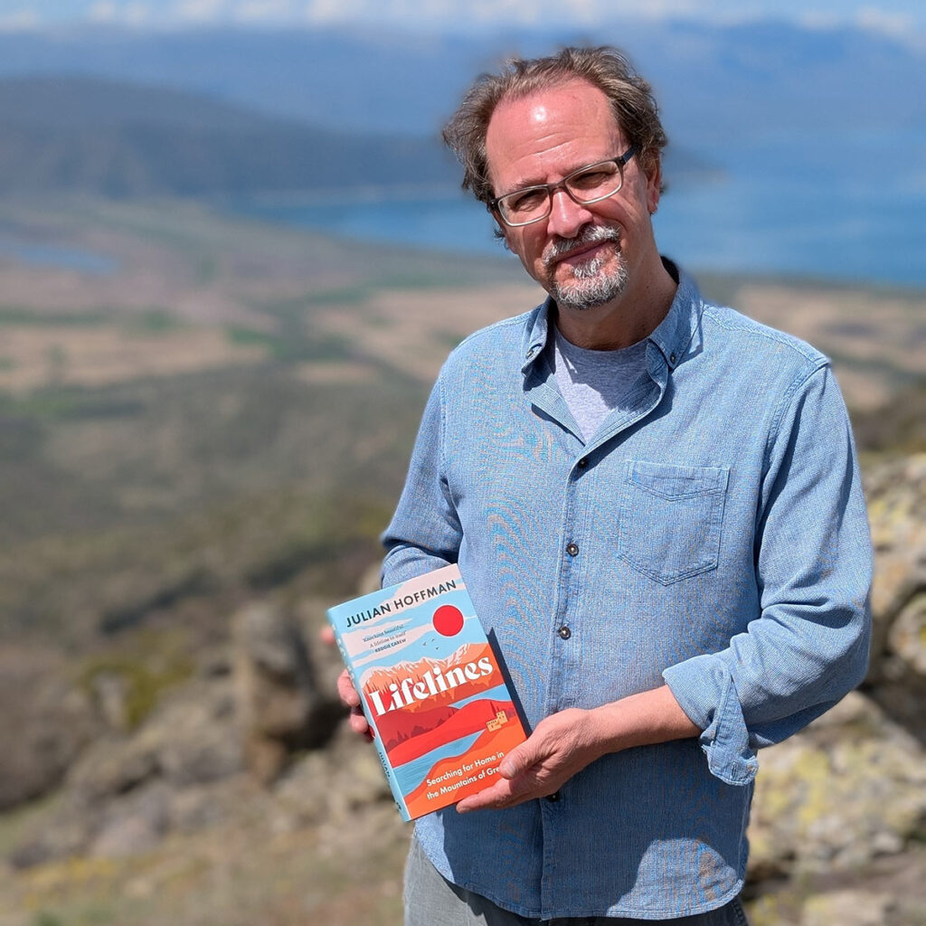 A man wearing a denim shirt holds a book up while standing outside.