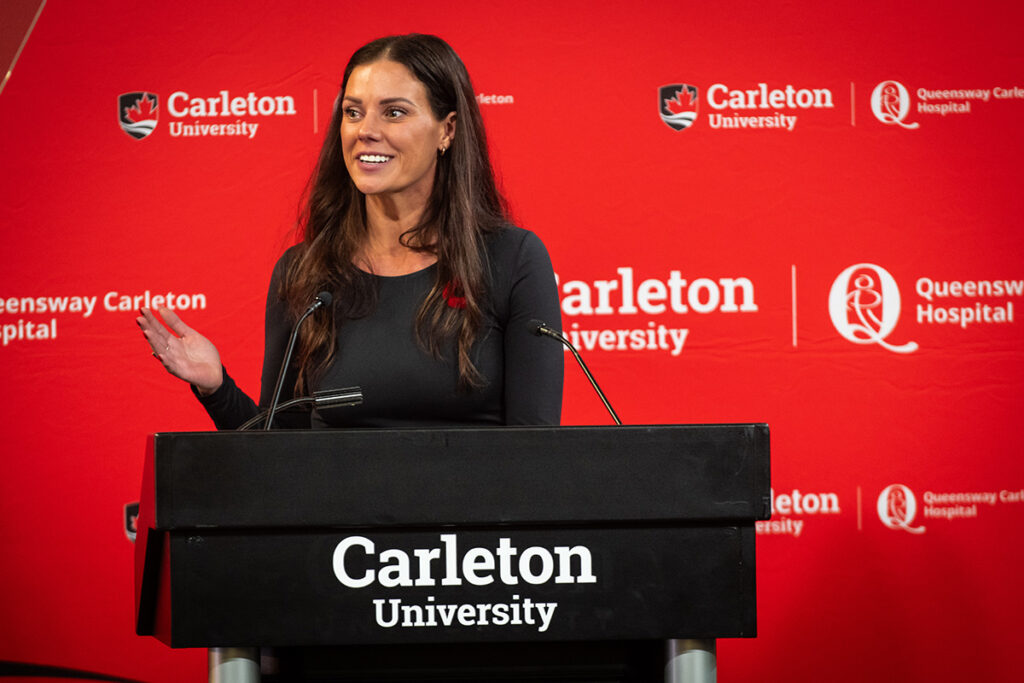 A woman addresses a crowd from a podium, with a red backdrop behind her.
