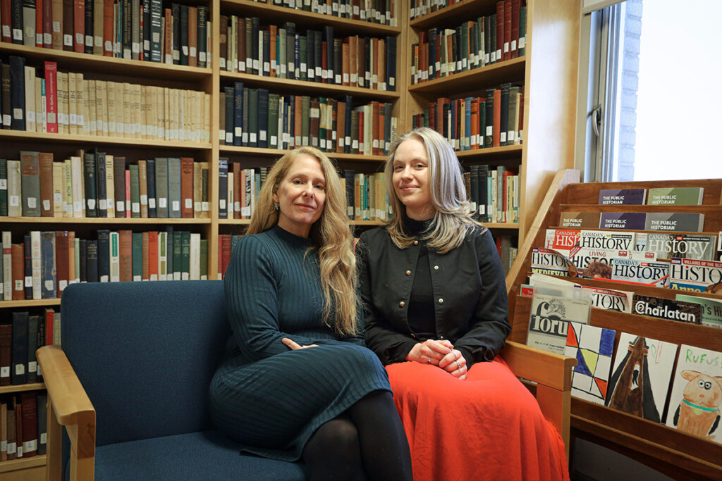 Two women posing for a photo while sitting on a sofa inside an office.