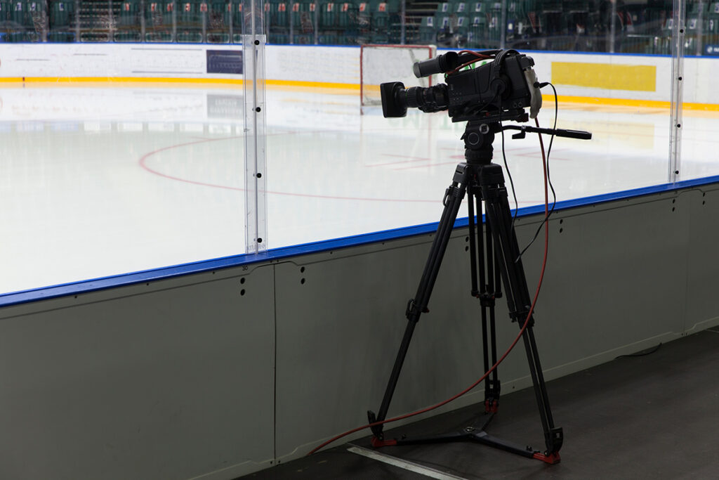 A professional video camera rests on a tripod, just outside of an ice rink.