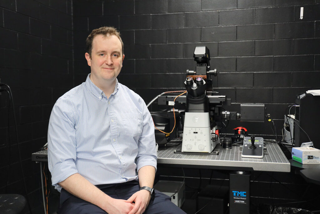 A man wearing a dress shirt poses for a picture while sitting next to a large microscope.