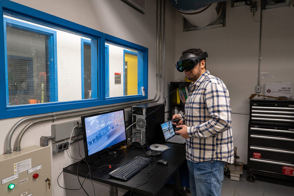 A man stands in front of a large computer monitor while wearing a VR headset.