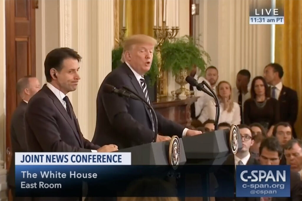 President Donald Trump speaks into a microphone while standing at a press conference podium.