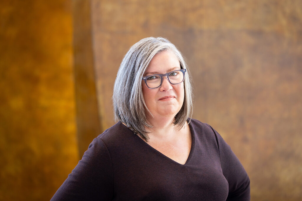 A woman with glasses, wearing a purple shirt, poses for a professional headshot.