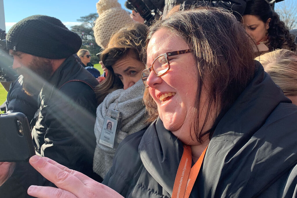 A woman wearing glasses records video with her mobile phone while standing in a large group of reporters.