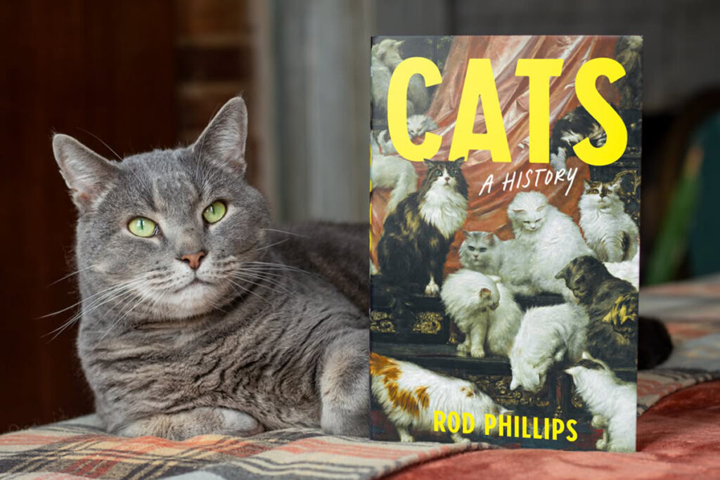 A cat sits next to the book Cats: A History by Rod Phillips.