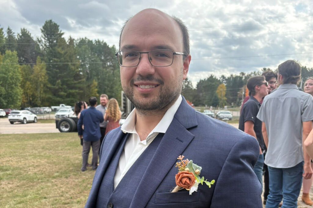 A man with glasses dressed in a blue suit smiles for the camera amongst a group of other people.
