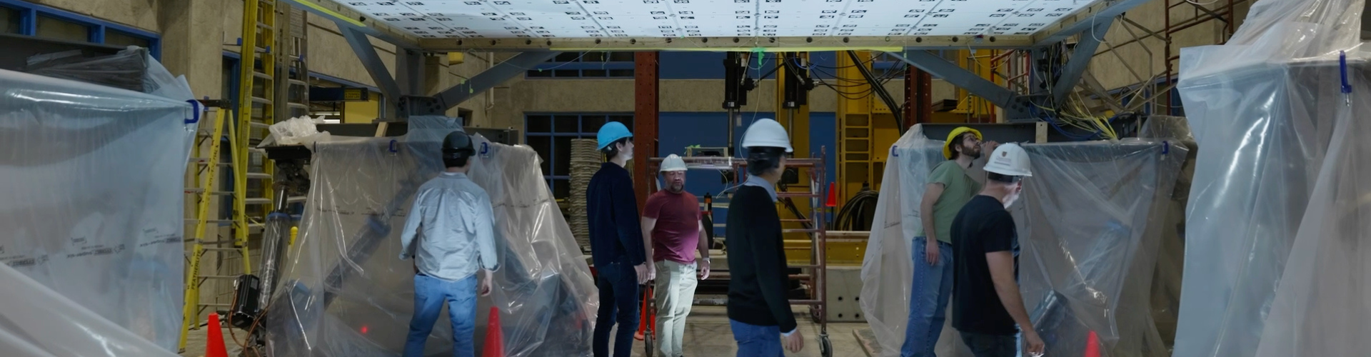 A group of people in hard hats and casual clothing stand in a construction site surrounded by plastic sheets and scaffolding, conveying a busy, industrious atmosphere.