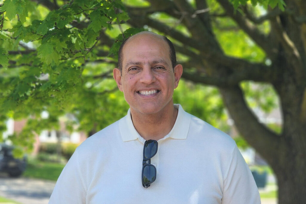 A man standing in front of a tree, smiles for the camera with sunglasses hanging from his shirt.