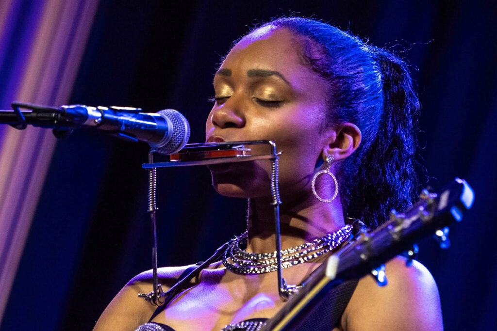 A woman sings into a microphone while playing a guitar and harmonica.