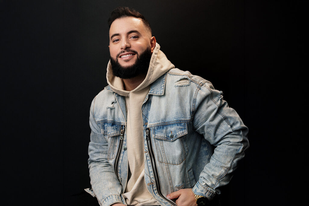 On a dark background, a man wearing a jean jacket over a beige hoodie, smiles for the camera.