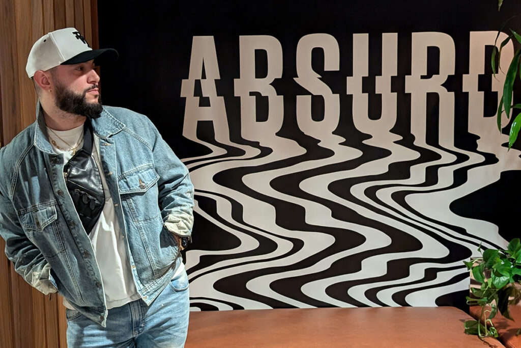 A man wearing a jeans and a jean jacket leans against a wall while looking at large Absurd sign.