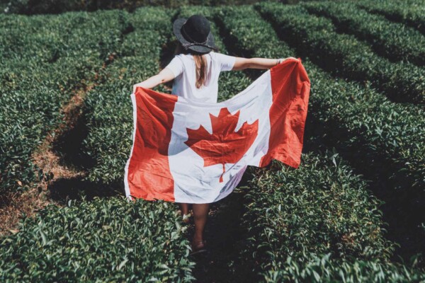 A woman in a hat carries a Canadian flag through a lush green field.