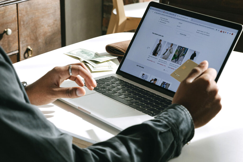A hand holds up a credit card, with an online store visible on the screen in front of them.